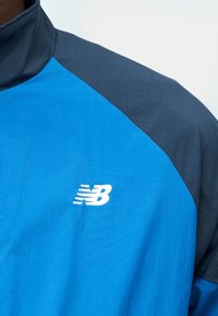 Μπλε και σκούρο γκρι αθλητικό μπουφάν με ψηλό γιακά, με λευκό λογότυπο New Balance στο στήθος.