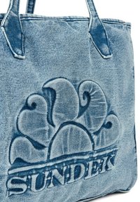 Sac en denim bleu clair avec un design floral en relief et le nom de la marque "SUNDEK". Il dispose de poignées robustes et d'un tissu texturé.