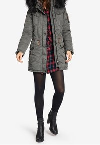 Vrouw in een groene parka met een bontkraag over een rood geruit jurkje, zwarte panty en zwarte enkellaarsjes, lopend tegen een witte achtergrond.