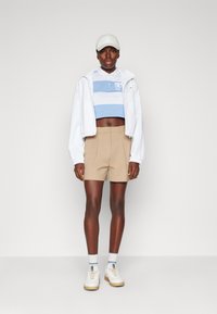Chaqueta blanca corta, polo a rayas en azul claro y blanco, pantalones cortos beige, zapatillas blancas y calcetines a rayas grises; fondo neutro.