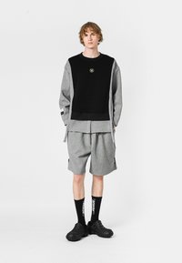 Sudadera gris y negra con un logo, combinada con pantalones cortos grises que presentan un detalle de cadena. El modelo lleva calcetines y zapatos negros.
