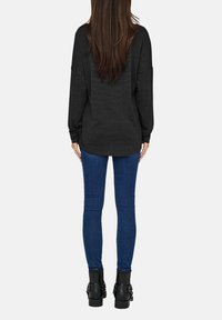 Jersey gris oscuro de gran tamaño y jeans ajustados azules. Botines negros con tiras. El cabello largo y liso cae sobre la parte posterior del jersey.