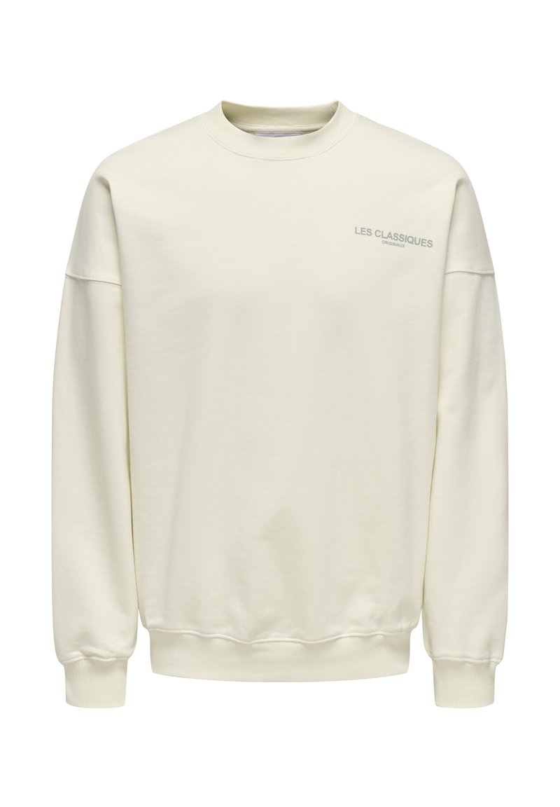 Cremfarbener Pullover aus weichem Stoff. Mit Rundhalsausschnitt, überschnittenen Schultern und kleinem blauen Text "LES CLASSIQUES" auf der Brust.