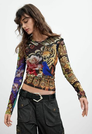 Donna con capelli lunghi castani che indossa una maglia colorata a maniche lunghe con motivi di tigre, rosa e uccello blu abbinata a pantaloni neri.