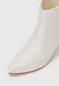 Bottine blanche à bout pointu avec une texture subtile de dentelle florale et une semelle lisse sur fond clair.