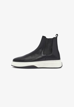 Botín Chelsea negro de cuero con paneles elásticos en los laterales, suela de goma blanca y un lazo en la parte trasera para facilitar su colocación. Textura suave y diseño aerodinámico.