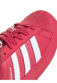 Närbild av röd sneakersko med vita zigzag-ränder, texturerad gummiskalnäsa och röda snören.