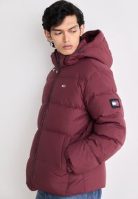 Burgundy puffer jacket with a hood, featuring quilted sections, a zip front, and a logo patch on the sleeve. Soft, insulated material.