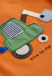 Appliqué d'un camion vert sur un fond en tissu orange, avec des détails brodés, une fenêtre rayée et le texte "il est temps de creuser !" en noir.