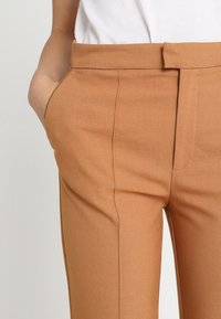 Brown tailored trousers with a smooth texture, high waist, and side pockets. The fabric appears lightweight and has a crease-free finish.
