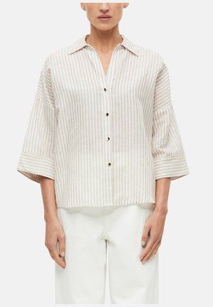 Femme portant une chemise à rayures verticales beige et blanche avec de larges manches et un pantalon blanc sur fond uni.