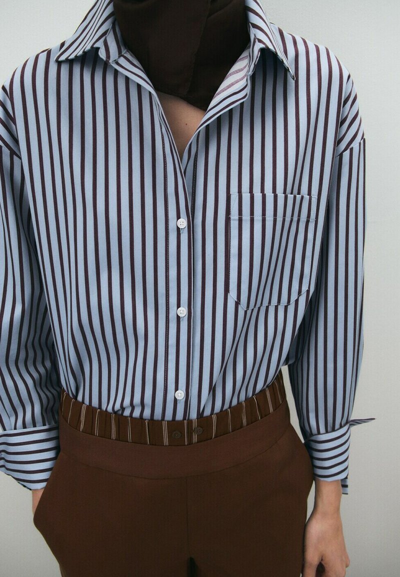 Chemise bleue à manches longues avec des rayures verticales marron, rentrée dans un pantalon marron taille haute avec une ceinture assortie à rayures.