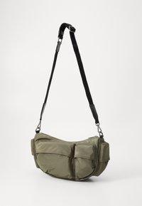 Sac en nylon vert de forme incurvée, doté d'une fermeture éclair, de plusieurs poches extérieures et d'une bandoulière noire réglable avec quincaillerie.