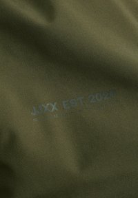 Tessuto verde oliva con testo stampato "JJXX EST. 2020" e slogan più piccolo "Sii audace, sii brillante e rimani dedicato."