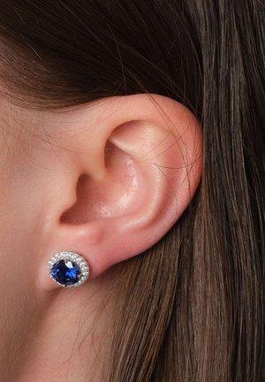 Runde Ohrringe haben einen tiefblauen Mittelstein, der von kleinen weißen Steinen umgeben ist, und sind aus silberfarbenem Metall gefertigt. Sie werden am Ohr einer Person getragen.