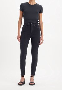 Jeans skinny de cintura alta negros combinados con una camiseta ajustada de color oscuro. El conjunto presenta un diseño recortado con lazos laterales y sandalias atadas.