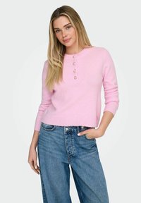 Pull rose en tricot avec un col rond, orné de cinq boutons en forme de cœur. Associé à un jean bleu ample, le mannequin se tient avec une main dans la poche.