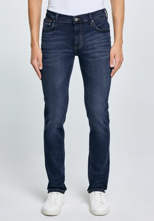 Mees tumedates sinistes kitsastes teksapükstes ja valgetes vabaajajalatsites, seisab käed külgedel lõdvestunult ühevärvilise tausta ees.