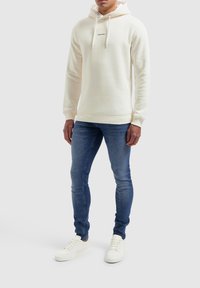 Crème-kleurige hoodie met een voorkeurszak, een capuchon met trekkoord en zwarte tekst "PURE PATH". Gecombineerd met blauwe jeans en witte sneakers.