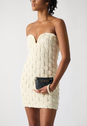 Robe crème sans bretelles avec un motif texturé en vagues et un décolleté en V. Mini sac Guess noir avec détail en logo doré tenu dans une main.