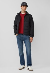 Junger Mann mit schwarzer Mütze, schwarzer gesteppter Jacke, rotem Pullover, blauen Jeans und weißen Sneakers, der vor einem einfarbigen Hintergrund steht.