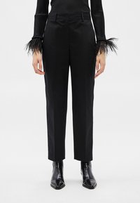 Persona, kas valkā melnas pieguļošas bikses, melnus potīšu zābakus un melnu garroku topu ar spalvu manšetēm, stāv uz baltas virsmas.