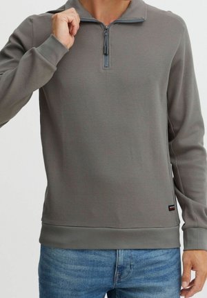 Homme portant un pull gris à manches longues avec une fermeture éclair jusqu'au col et un jean bleu, tenant la fermeture éclair près du col, sur un fond uni.