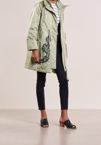 Parka imbottita verde con cappuccio, caratterizzata da un'applicazione floreale sul lato. Abbinata a jeans neri e mule aperte nere con tacchi in legno.