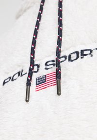 Felpa con cappuccio in pile bianco con scritta nera "POLO SPORT", cordini decorati a motivi rossi, bianchi e blu, e piccolo dettaglio ricamato con bandiera americana.