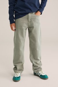 Gai pelēki džinsu bikses ar brīvu piegriezumu, ar sānu kabatām un taisnām kājām, savienotas ar zaļiem un baltiem kedām.