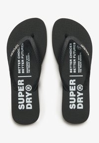 Tongs noires avec une semelle texturée et des straps en caoutchouc. Présente un texte imprimé blanc sur la semelle intérieure et une marque sur les straps.