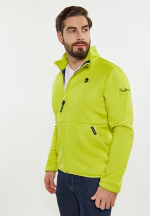 Jasno limonkowy polar z wysokim kołnierzem, zamek błyskawiczny, boczne kieszenie oraz detal z logo, zestawiony z ciemnoniebieskimi dżinsami.