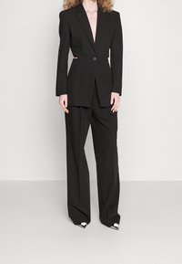 Completo nero su misura con un blazer strutturato a profondo scollo e vita aderente, abbinato a pantaloni larghi e scarpe con punta.