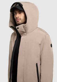 Chaqueta beige con capucha, fabricada con un tejido suave y resistente al agua, con cremallera frontal y bolsillos laterales. Presenta un logo negro en la manga.