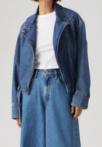 Tummansinisestä denim-takista, joka on lyhytmallinen, on terävä kaulus, edessä vetoketjukiinnitys ja kaksi sivutaskua. Yhdistetty vaaleansinisiin leveisiin farkkuihin.