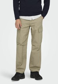 Beiget sotilashousut, joissa on useita taskuja, suoralahkeinen malli ja ommellut yksityiskohdat. Yhdistetty tummansiniseen neuleeseen, joka on puettu valkoisen kauluksettomaan paitaan päälle.