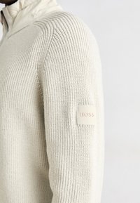 Pull en tricot beige clair avec col montant, présentant une texture côtelée et un patch de marque sur la manche gauche.