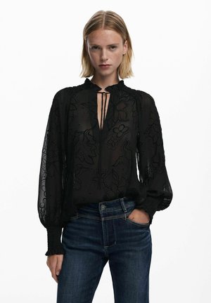Blusa nera trasparente con ricami floreali, maniche a palloncino lunghe e scollatura arricciata; indossata con jeans in denim scuri.