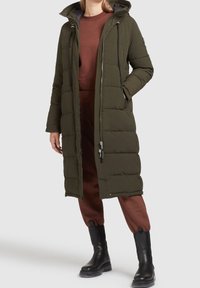 Lang, olivengrøn pufferjakke med hætte, der har lynlås lukning, sidelommer og et quiltet mønster. Parret med et brunt outfit og sorte støvler.