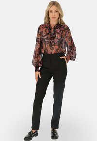 Przezroczysta czarna bluzka z kolorowym wzorem paisley, wysoki kołnierz z wstążką, zestawiona z dopasowanymi czarnymi spodniami i błyszczącymi czarnymi butami.