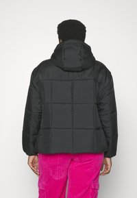 Veste rembourrée noire avec capuche, présentant un design matelassé et une texture lisse. Portée sur un pantalon en velours côtelé rose, affichant un style décontracté.