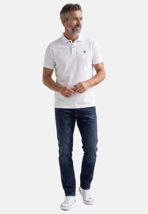 Mann mit grauen Haaren und Bart, der ein weißes Poloshirt, dunkelblaue Jeans und weiße Turnschuhe trägt, steht mit verschränkten Händen.