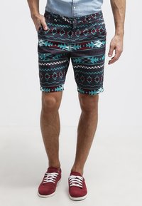 Muž v šortkách s modrými, vínovými a černými geometrickými vzory, světle modré košili a červených teniskách s bílými tkaničkami.