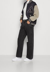 Svarta denimbyxor med vida ben, vita sneakers, en marinblå och beige collegejacka med ett broderat bokstav och randiga ärmslut, samt en vit polotröja.