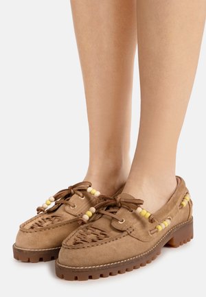 Mocassins en daim marron avec semelles en caoutchouc épaisses, perles décoratives jaunes et beiges, lacets à l'avant, portés pieds nus.