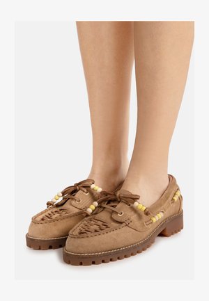 Mocassins en daim marron avec semelles en caoutchouc épaisses, perles décoratives jaunes et beiges, lacets à l'avant, portés pieds nus.