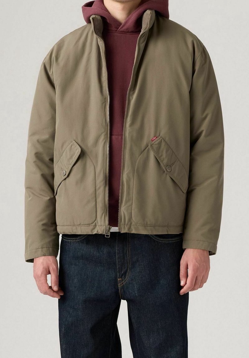 Homme portant une veste zippée vert olive par-dessus un sweat à capuche bordeaux et un jean bleu foncé, debout les mains détendues le long du corps.