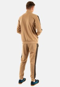 Uomo in piedi di spalle che indossa un tuta color sabbia con strisce nere e sneakers blu con suola in gomma su sfondo bianco.