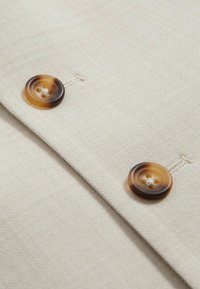 To brune gradientknapper syet på beige tekstureret stof med synlige knaphuller og overlappende stoflag.