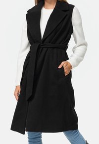 Manteau sans manches noir avec une ceinture, en matériau doux semblable à de la laine, avec un large col et deux poches avant.